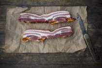 Slices of streaky bacon on parchment with knife on dark wood — Stock Photo