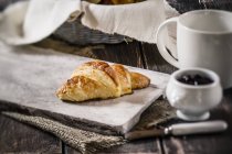 Croissant en plato de madera, cesta con croissants, taza de café y olla con mermelada - foto de stock