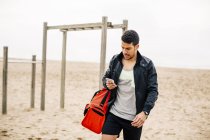 Young sporty man using cell phone on sandy beach — Stock Photo