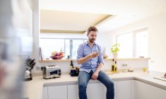 Hombre sentado en la cocina usando un teléfono inteligente - foto de stock