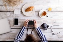 Uomo che lavora a tavola con croissant e tè, guardando la sua macchina fotografica — Foto stock