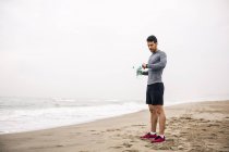 Спортивный молодой человек с бутылкой на пляже проверяет часы — стоковое фото