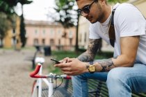 Молодой человек сидит на скамейке и смотрит на мобильный телефон — стоковое фото