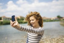 Italia, Verona, retrato de una mujer sonriente tomando selfie con smartphone a orillas del río - foto de stock