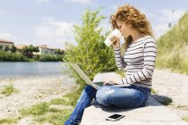 Femme boire du café pour aller tout en utilisant un ordinateur portable au bord de la rivière — Photo de stock