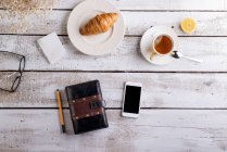 Tavolo con croissant, tè, smartphone e organizzatore personale — Foto stock