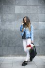 Jeune femme sur téléphone portable avec un bouquet de fleurs dans un sac contre un mur gris — Photo de stock