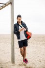 Молодой спортсмен пользуется мобильным телефоном на песчаном пляже — стоковое фото