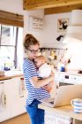Mutter mit Baby in Küche schaut auf Laptop — Stockfoto