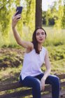 Mujer tomando una selfie mientras está sentada en un banco en el parque - foto de stock
