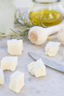 Diced sheep cheese, glass of olive oil, garlic bulbs, rosemary and knife on white marble — Stock Photo