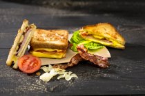 Gros plan de sandwich au fromage, jambon, bacon, tomates et salade sur une surface en bois noir — Photo de stock