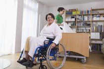 Mujer mayor sentada en silla de ruedas otra mujer arreglando cama médica - foto de stock