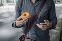 Imagen recortada de empresario sosteniendo donut y teléfono inteligente, Vista parcial - foto de stock