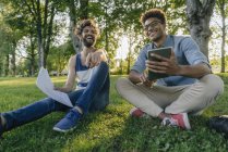 Zwei fröhliche multikulturelle Freunde sitzen mit Handy und Papieren im Park — Stockfoto