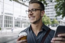 Ritratto di giovane uomo d'affari ridente con caffè da portare via e smartphone — Foto stock
