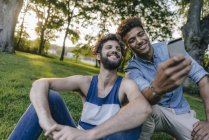 Dos amigos multiculturales felices compartiendo el teléfono celular en el parque - foto de stock
