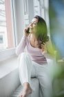 Portrait of laughing young woman on the phone sitting on heater in loft looking out of window — Stock Photo
