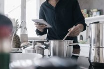Man standing in kitchen and reading news on his digital tablet while cooking — Stock Photo