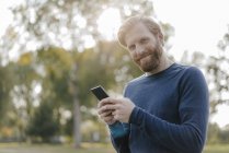 Портрет улыбающегося мужчины с сотовым телефоном в парке, смотрящего в камеру — стоковое фото