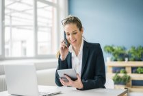 Businesswoman sitting at desk, talking on the phone, looking at digital tablet — Stock Photo