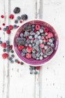 Bowl of deep frozen red currents, raspberries, blueberries and blackberries on white wood — Stock Photo