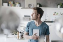 Человек с планшетом стоит на кухне и смотрит на расстояние — стоковое фото
