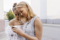 Two smiling young women sharing cell phone on street — Stock Photo