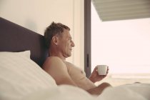 Расслабленный мужчина в постели держа чашку кофе — стоковое фото