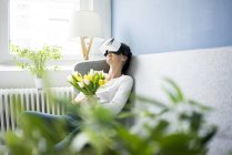 Smiling woman sitting on couch with VR glasses, holding bunch of tulips — Stock Photo