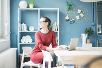 Mulher sorridente sentada à mesa com laptop e olhando pela janela — Fotografia de Stock