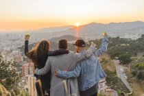 Espanha, Barcelona, três amigos com garrafas de cerveja abraçando em uma colina com vista para a cidade ao pôr do sol — Fotografia de Stock