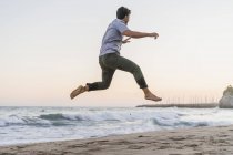 Щасливий молодий чоловік стрибає в повітрі на пляжі на заході сонця — стокове фото
