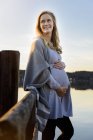 Sorridente donna incinta in piedi sulla riva del lago — Foto stock