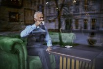 Элегантный пожилой мужчина сидит на диване в кафе и пьет кофе — стоковое фото