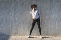 Сміється жінка в капелюсі, стоячи перед плиткою стіни на сонячному світлі — стокове фото