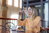 Smiling young woman in a cafe taking cell phone picture — Stock Photo