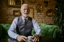 Портрет элегантного пожилого человека, сидящего на диване в кафе и улыбающегося — стоковое фото