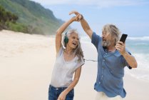 Bonito casal sênior com fones de ouvido dançando na praia — Fotografia de Stock