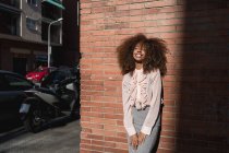 Portrait of smiling young woman with afro hairdo leaning against brick wall in the city — Stock Photo