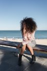Beautiful young woman with afro hairdo sitting on a bench at the beach — Stock Photo