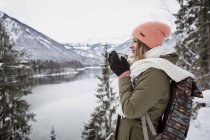 Junge Frau mit Heißgetränk steht in alpiner Winterlandschaft mit See — Stockfoto
