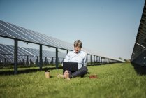 Mature man sitting barefoot on meadow, laptop and coffee to go cup, solar plant — Stock Photo