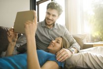 Casal sorrindo olhando para tablet e relaxando no sofá em casa — Fotografia de Stock