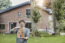 Retrato del hombre en el jardín de su casa sosteniendo modelo de la casa con la mujer en el fondo que miente en la hamaca - foto de stock