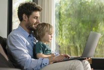 Père et fils regardant ordinateur portable sur le canapé à la maison — Photo de stock