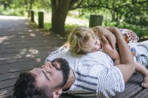 Kleines Mädchen umarmt seinen Vater, der auf einem Holzsteg in der Natur liegt — Stockfoto