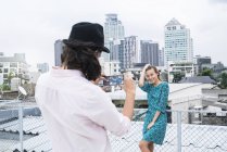 TJoven con smartphone tomando fotos de su novia posando en la azotea - foto de stock