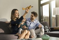 Padres felices e hijo jugando con el avión de juguete de madera en el sofá en casa - foto de stock