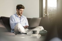 Hombre sentado en el sofá en casa usando el ordenador portátil - foto de stock